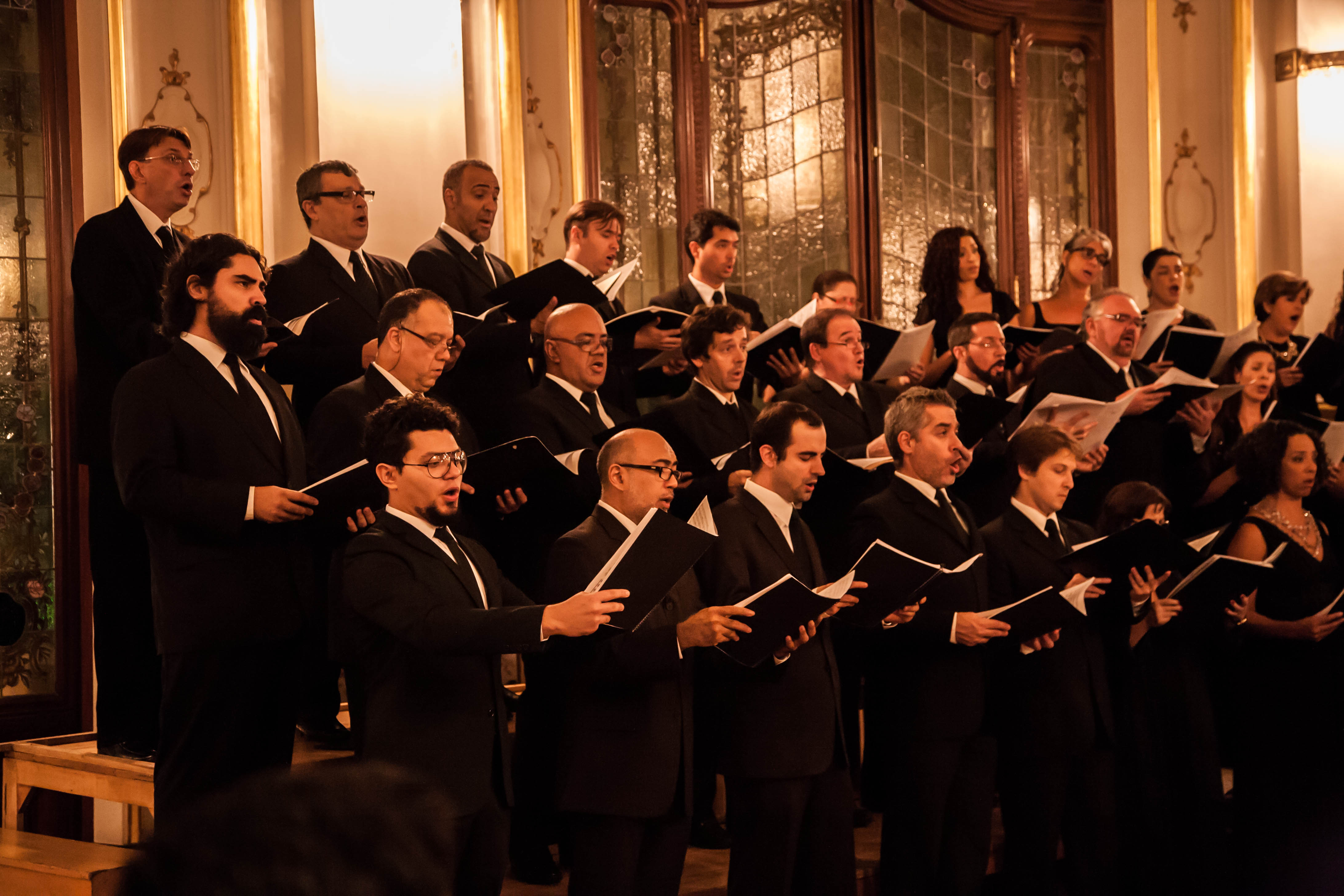 Na imagem cantores do coral vestem preto e estão segurando o repertório nas mãos