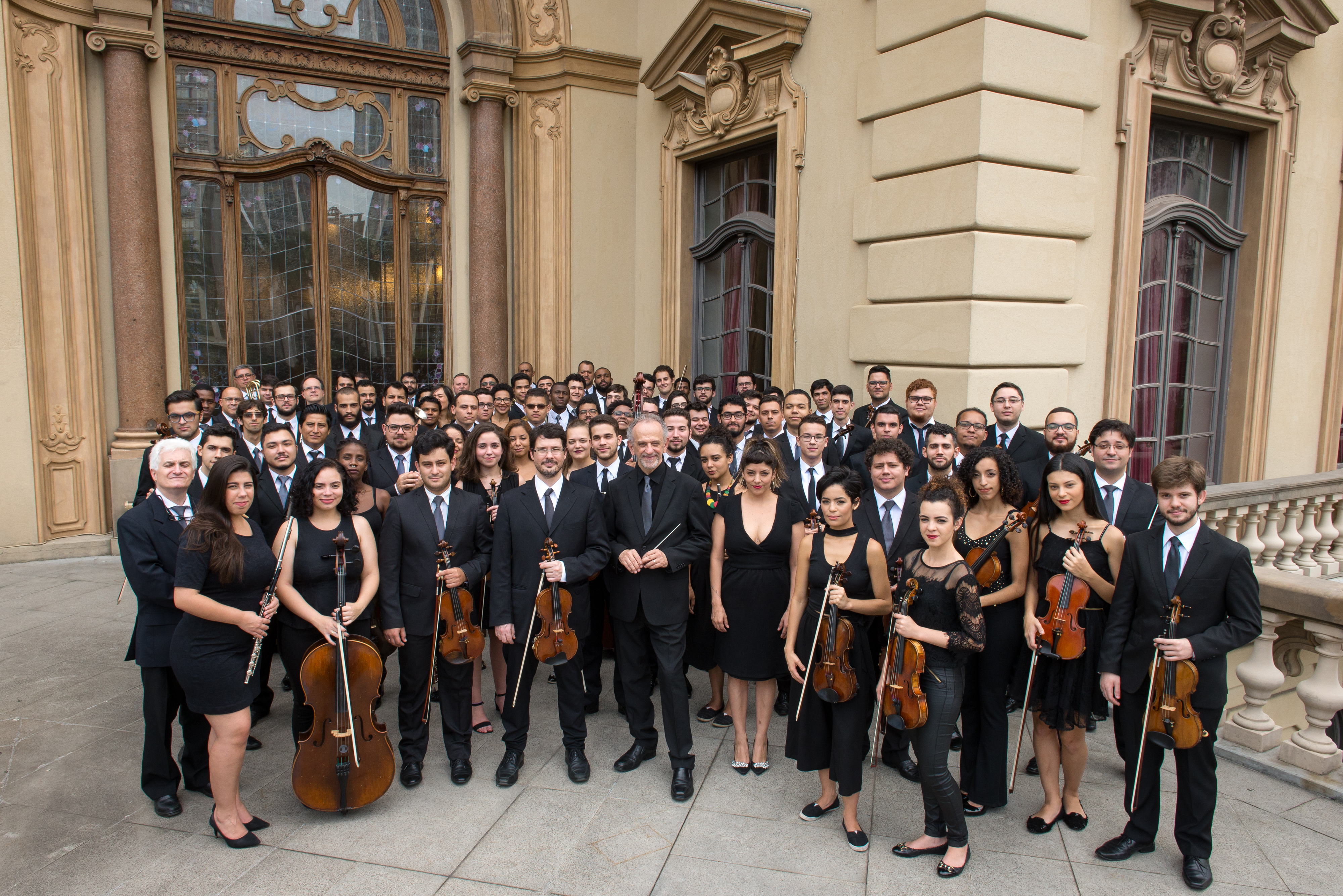 Uma foto com uma orquestra completa na fachada de um prédio, todos os músicos estão de preto com seus instrumentos em mãos.