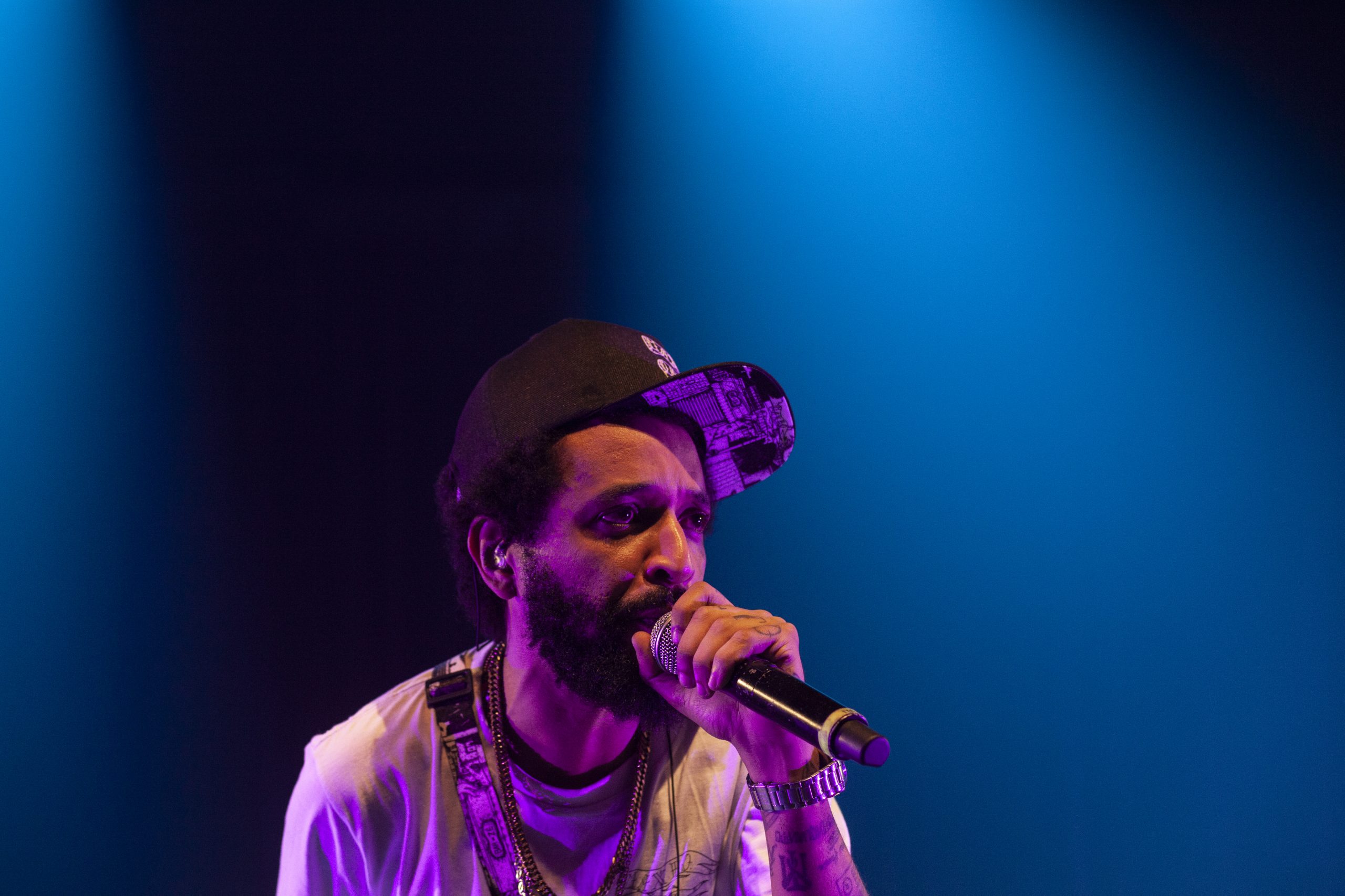 MC Max B.O. com microfone próximo a boca cantando. Ele usa um boné e uma camiseta branca